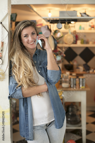 Fototapeta Naklejka Na Ścianę i Meble -  Beautiful young woman in a hipster kitchen talks to her phone dressed in a jeans and a white shirt