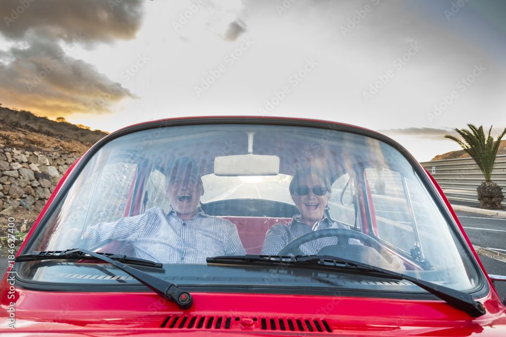 Elderly couple with hat, with glasses, with gray and white hair, with casual shirt, on vintage red car on vacation enjoying time and life. With a cheerful mobile phone smiling