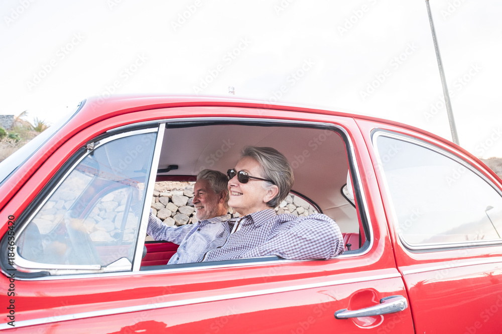 Elderly couple with hat, with glasses, with gray and white hair, with casual shirt, on vintage red car on vacation enjoying time and life. With a cheerful mobile phone smiling Elderly couple with hat, with glasses, with gray and white hair, with casual shirt, on vintage red car on vacation enjoying time and life. With a cheerful mobile phone smiling