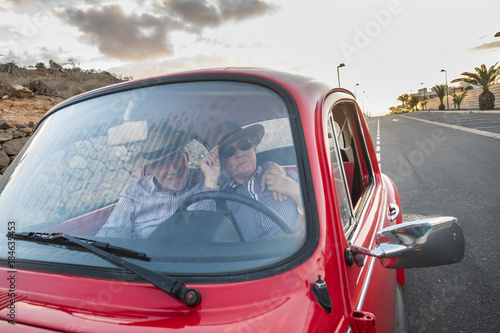 Elderly couple with hat, with glasses, with gray and white hair, with casual shirt, on vintage red car on vacation enjoying time and life. With a cheerful mobile phone smiling 