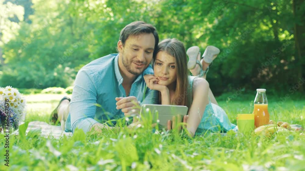 Romantic couple lying on the grass at the picnic in the picturesque green park and watching something on the tablet computer. Sunny day. Portrait shot. Outdoors Romantic couple lying on the grass at the picnic in the picturesque green park and watching something on the tablet computer. Sunny day. Portrait shot. Outdoors