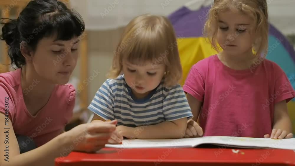 The educator and two little children girl and boy sitting at table drawing. Brother and sister and their mother drawing with colorful crayons and markers The educator and two little children girl and boy sitting at table drawing. Brother and sister and their mother drawing with colorful crayons and markers
