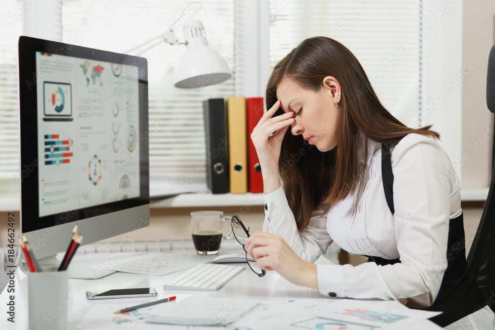 Beautiful tired perplexed and stress brown-hair business woman in suit and glasses sitting at the desk with cup of coffee, working at contemporary computer with documents in light office Beautiful tired perplexed and stress brown-hair business woman in suit and glasses sitting at the desk with cup of coffee, working at contemporary computer with documents in light office