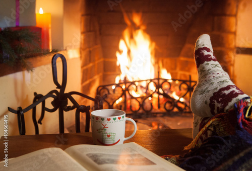 A woman in warm knitted socks with a cup of hot mulled wine and an old book in front of the fireplace. Evening fairy tales. Fantasy.