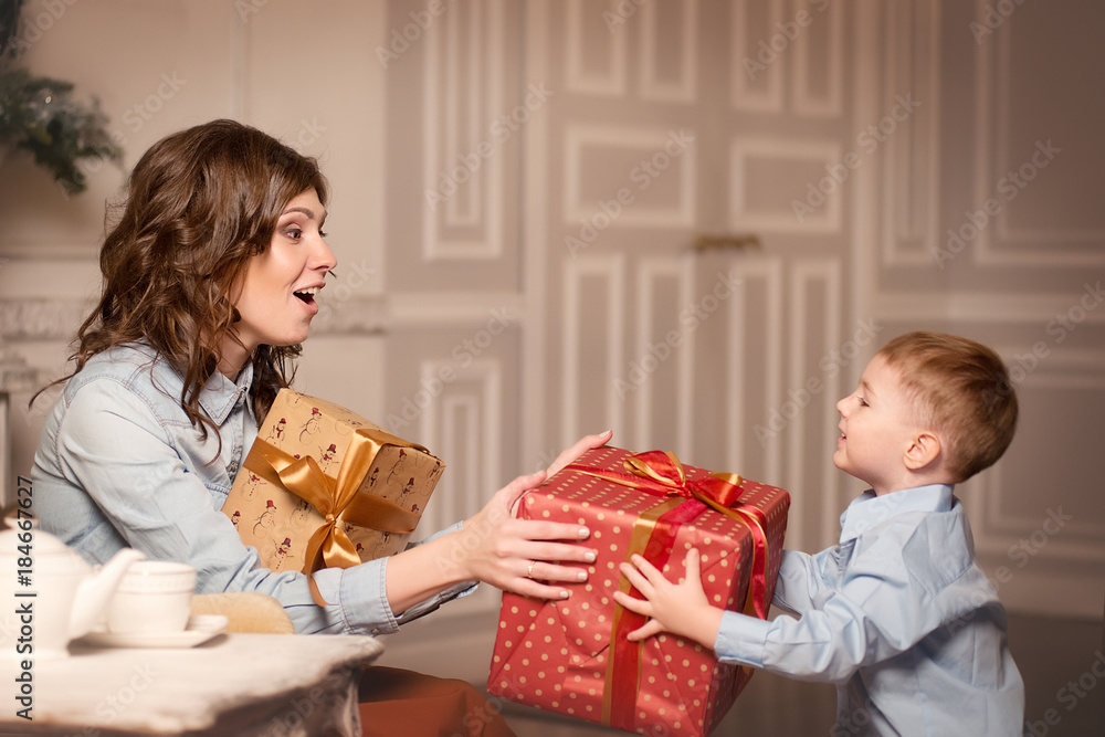 A young mother with an emotion of surprise receives a Christmas gift from his son vintage beige interiors A young mother with an emotion of surprise receives a Christmas gift from his son vintage beige interiors