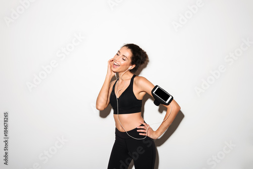 Pleased curly brunette fitness woman listening music by smartphone