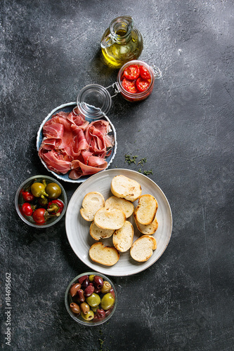 Ingredients for making tapas or bruschetta. Crusty bread, ham prosciutto, sun dried tomatoes, olive oil, olives, pepper, greens on plates over dark texture background. Top view with space