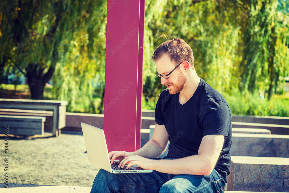Working Anywhere. Technology in our daily life. Wearing black T shirt, jeans, glasses, a young man with beard, mustache sitting at park, reading, typing, working on laptop computer. Filtered effect.. Working Anywhere. Technology in our daily life. Wearing black T shirt, jeans, glasses, a young man with beard, mustache sitting at park, reading, typing, working on laptop computer. Filtered effect..