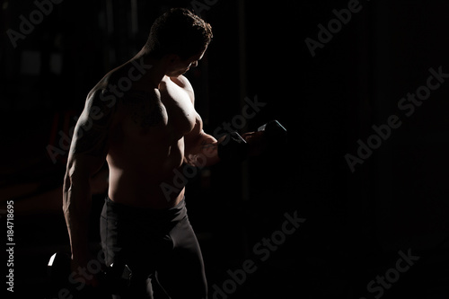 Handsome power athletic guy bodybuilder doing exercises with dumbbell. Fitness muscular body on dark background. © Artem Zakharov
