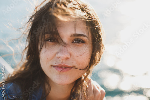 Beautiful young woman at the ocean standing against a turquoise blue sea with her hair blowing in the breeze