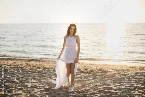 Beautiful girl bride in a white dress and sneakers, at sunset walks along the beach.