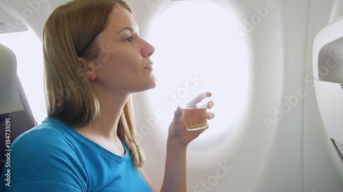 Woman flying in airplane in daytime. Thirsty female drinking white wine near window during flight turbulence photo