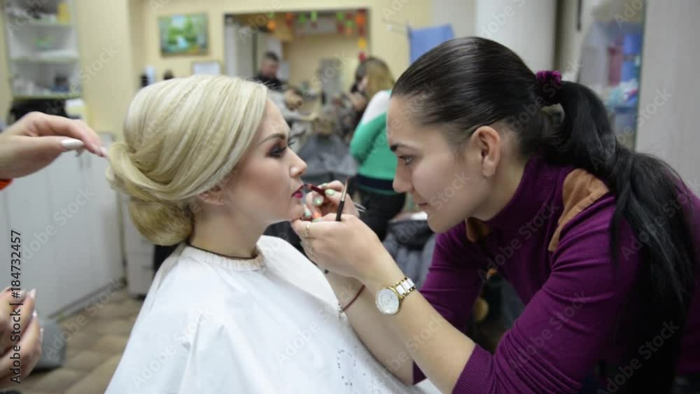 One visagiste makes hairstyle and other paints lips to girl model. Beauty salon - woman doing hair styling and makeup One visagiste makes hairstyle and other paints lips to girl model. Beauty salon - woman doing hair styling and makeup