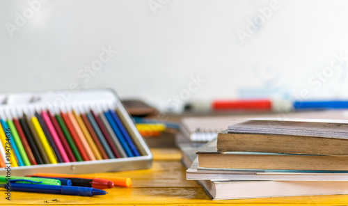 Coloring pencils, pens and closed books and notebooks on top of a yellow wooden table, with a white school board in the background