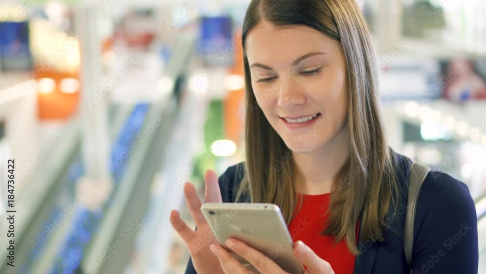 Young beautiful woman standing in shopping mall smiling. Using her smartphone, talking with friends. Shopping consumerism concept Young beautiful woman standing in shopping mall smiling. Using her smartphone, talking with friends. Shopping consumerism concept