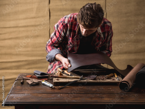 Working process of the leather belt in the leather workshop. Man holding crafting tool and working. Tanner in old tannery. Wooden table background. Warm Light for text and design.