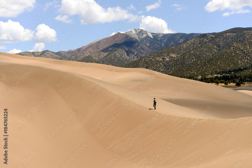 Hiking on Sand Dunes - A tourist is hiking on a sand ridge on a spring day at Great Sand Dunes National Park, Colorado, USA. The park sits at west base of Sangre de Cristo Range in San Luis Valley. Hiking on Sand Dunes - A tourist is hiking on a sand ridge on a spring day at Great Sand Dunes National Park, Colorado, USA. The park sits at west base of Sangre de Cristo Range in San Luis Valley.