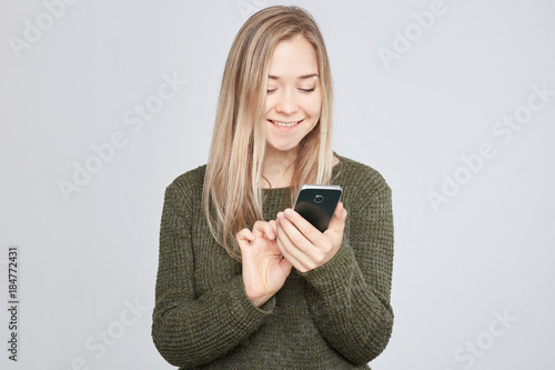 .Portrait of pretty young blond Caucasian woman, typing a message to her boyfriend on mobile phone, holding the gadget with one hand, wearing warm wool sweater, looking happy on white wall background.