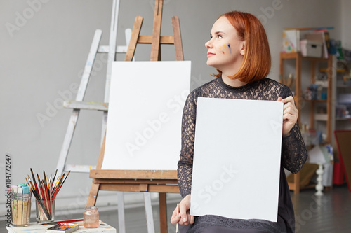 Portrait of prifile of redhead authentic female face having an empty vertical canvas mockup in hands with space for your advertising. Authentic lady painter sitting indoors in studio creating artworks photo