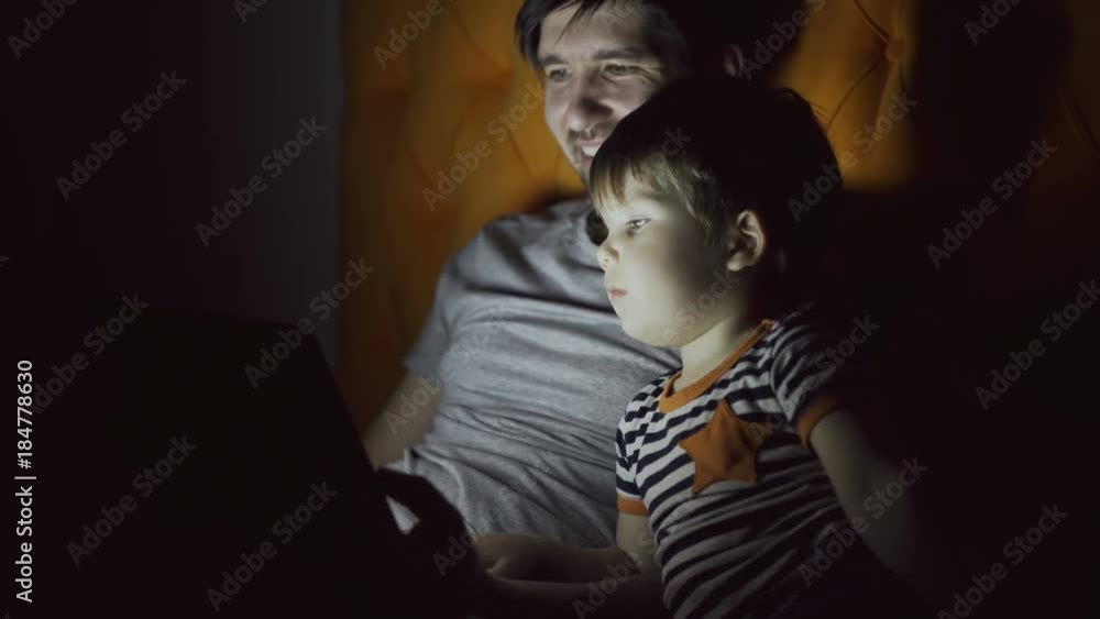 Young father and his little son watching cartoon movie using tablet computer and talking while lying in bed in evening at home Young father and his little son watching cartoon movie using tablet computer and talking while lying in bed in evening at home