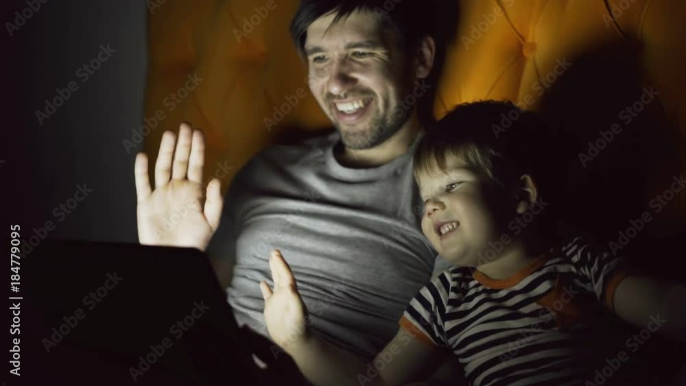 Loving father and his little son having online video chat with grandparents using tablet computer and while lying in bed in evening at home Loving father and his little son having online video chat with grandparents using tablet computer and while lying in bed in evening at home