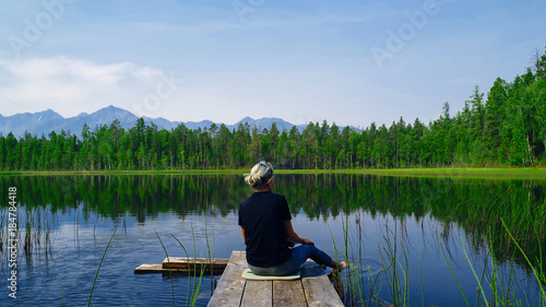 Lonely dreaming woman sitting on a wooden bridge pier looking at the mountains reflected in the mirror water of the forest lake. Serenity concept. Pure nature.