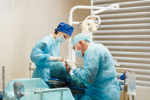 Male old professional dentist surgeon in uniform and female assistant helps to performing operation install dental implant teeth of woman patient in clinic light office with modern tools equipment.