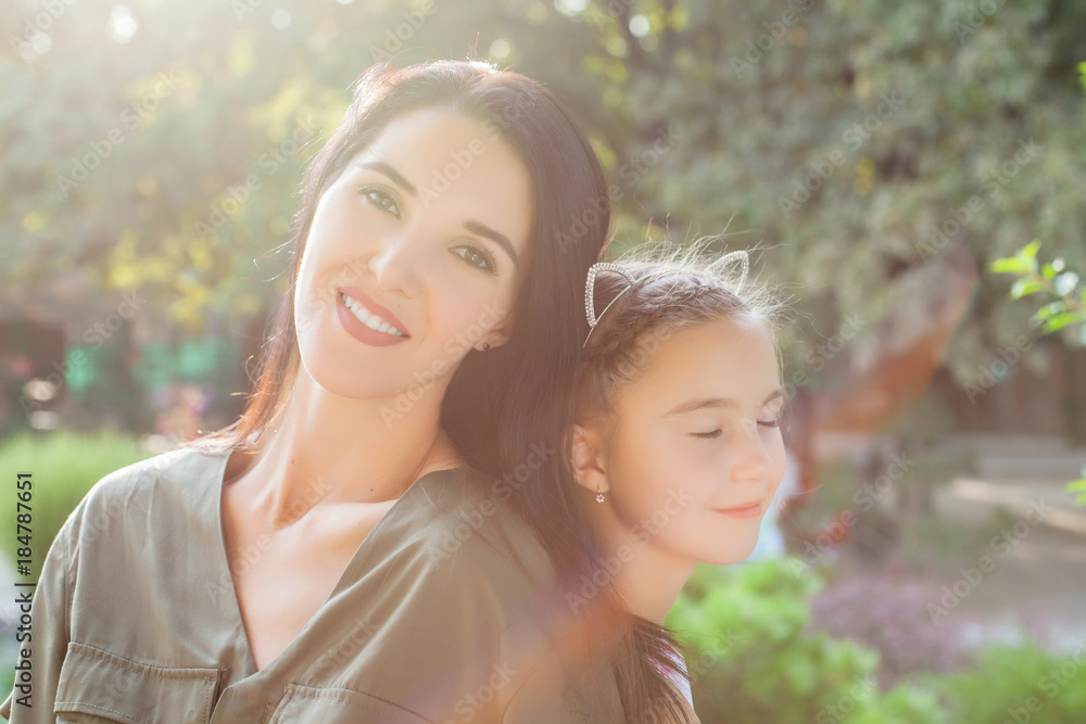 Young mother sitting with back to back with cute girl in beautiful sunlight in beautiful nature. Concept of happy family Young mother sitting with back to back with cute girl in beautiful sunlight in beautiful nature. Concept of happy family