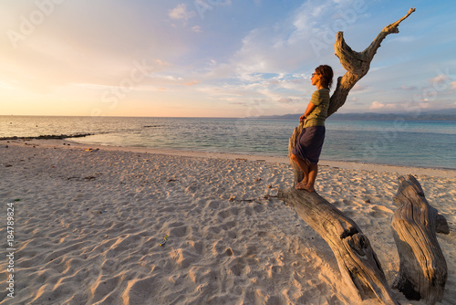 Woman resting on skeletal tree and watching a romantic colorful sunset on the beach of Tanjun Karang, Central Sulawesi, Indonesia. Wide angle shot, long exposure, blurred motion.