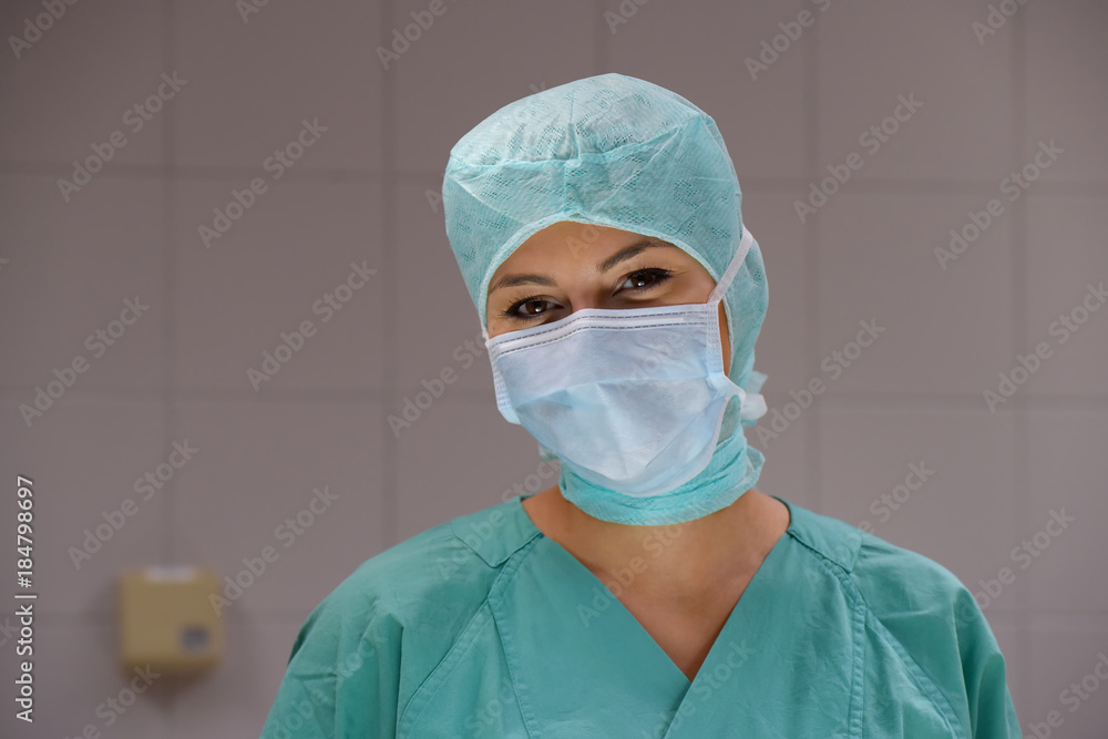 A young woman poses  in an operation theater  fully dressed as a theater nurse with a face mask  and green sterile medical work clothing.