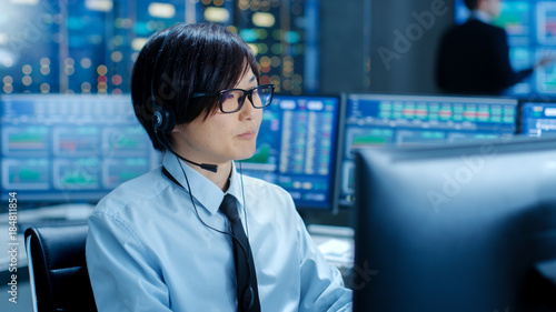 In the Network Operations Center Trader Makes Personal Client Call with a Headset. He's Surrounded by Monitors Showing Stocks Data and Graphs.