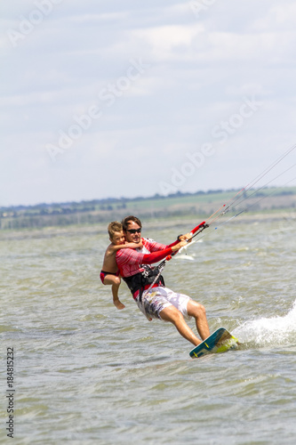 Dad and son are kitesurfing on the sea. The son clings to Papa's back
