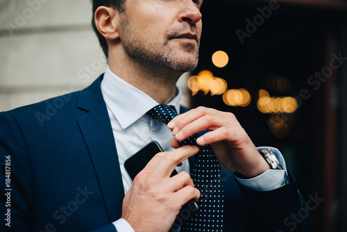 Midsection of businessman adjusting necktie while holding mobile phone photo