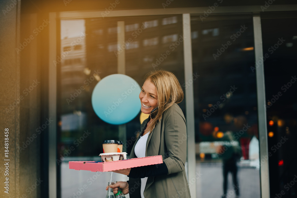 Smiling mature businesswoman talking on smart phone while holding food and drink against cafe in city Smiling mature businesswoman talking on smart phone while holding food and drink against cafe in city
