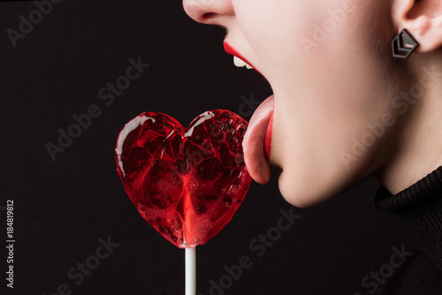 cropped image of woman licking heart shaped lollipop isolated on black