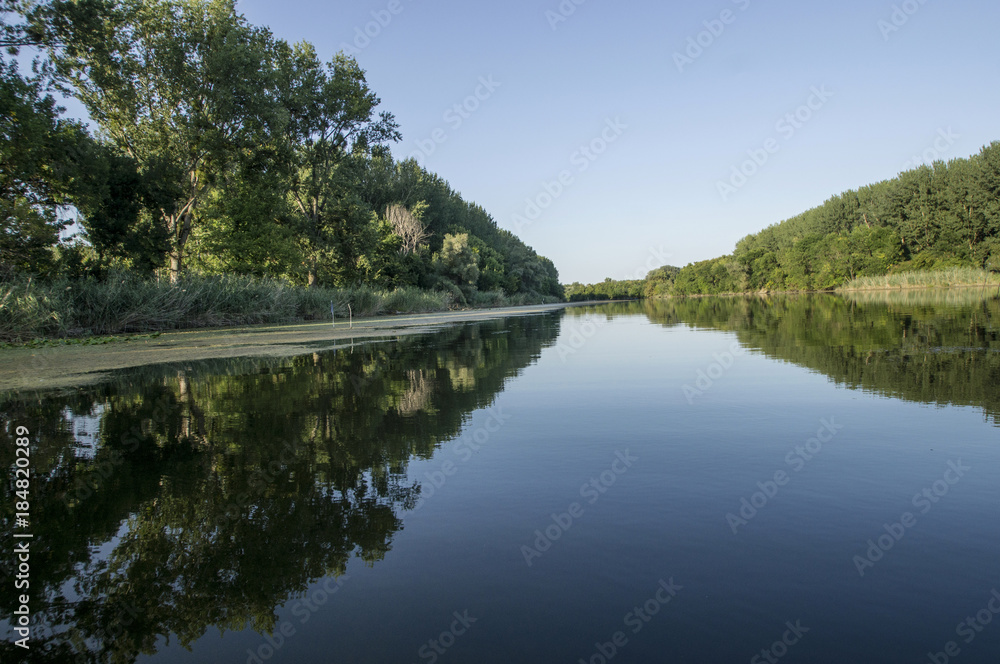 The  surface of the river acts as a mirror. The shores of the river are covered with lush vegetation. Trees and reeds are reflected on the surface of the river. On the horizon is the end of the trip. 