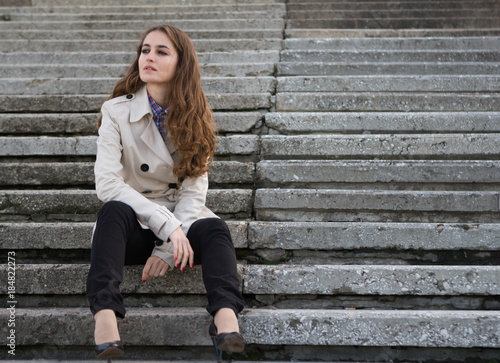 Young beautiful woman wearing beige jacket sitting on concrete stairs and looking away