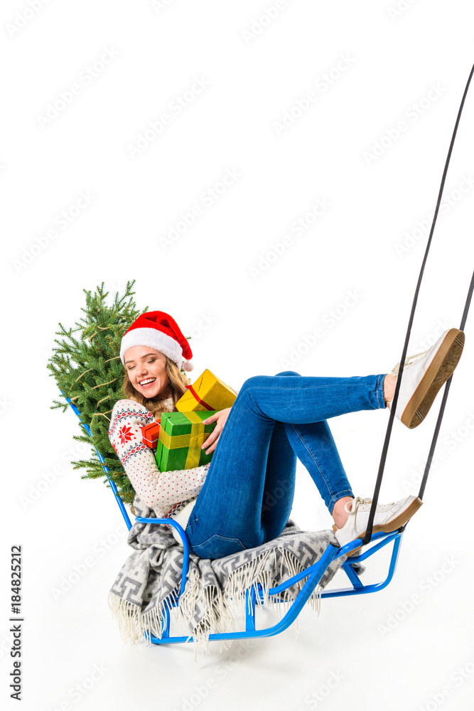 Obraz premium laughing girl in santa hat sitting on sled with gifts and christmas tree, isolated on white