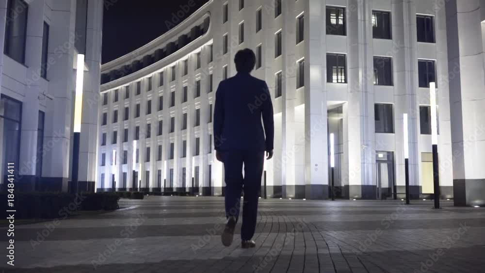 Rear view of a young businessman wearing a black suit and walking in a night city. Concept of success. Tracking real time establishing shot Rear view of a young businessman wearing a black suit and walking in a night city. Concept of success. Tracking real time establishing shot