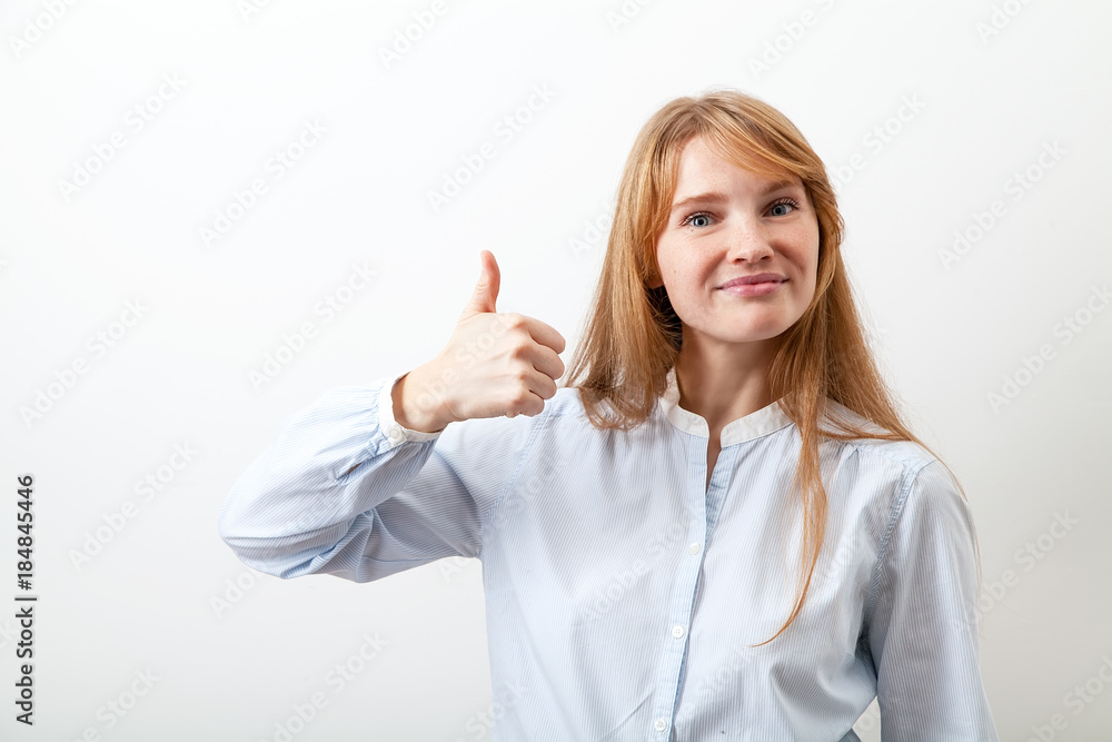 Studio portrait of pretty red-headed lady dressed in casual blue shirt smiling and showing big thumb up feeling enthusiastic and satisfied with a good quality. White background. Studio portrait of pretty red-headed lady dressed in casual blue shirt smiling and showing big thumb up feeling enthusiastic and satisfied with a good quality. White background.