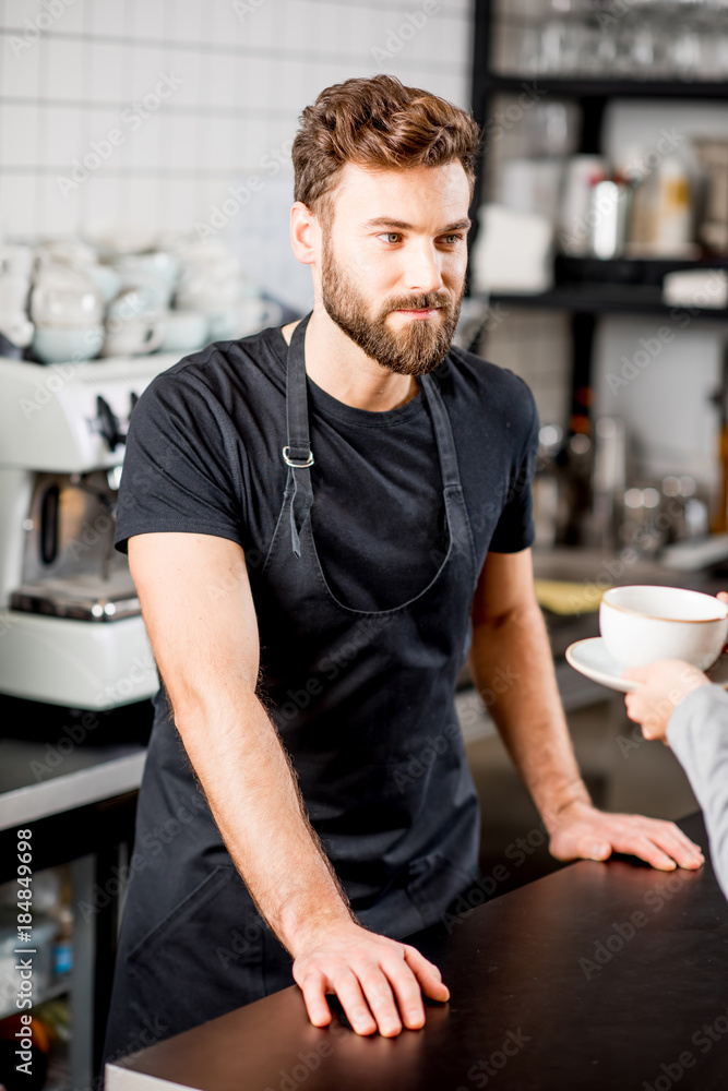 Portrait of a handsome barista in black t-shirt and apron standing at the bar of the modern cafe Portrait of a handsome barista in black t-shirt and apron standing at the bar of the modern cafe