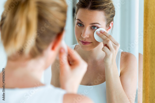 Beautiful young woman is cleaning her face while looking in the mirror in the bathroom.