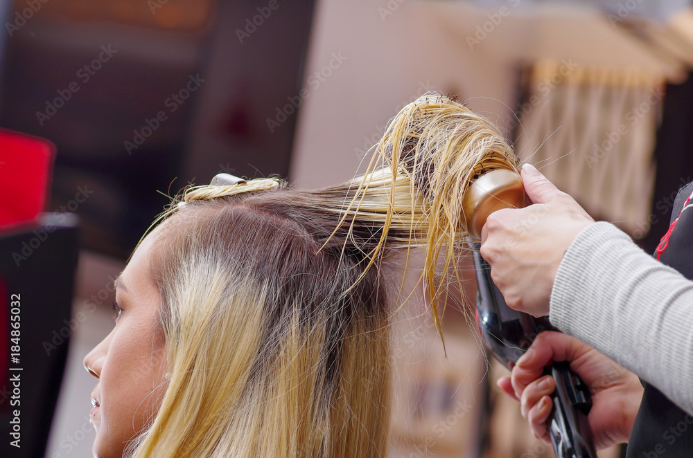 Fototapeta premium Close up of a hair stylist drying blond hair with hair dryer and round brush in a blurred background