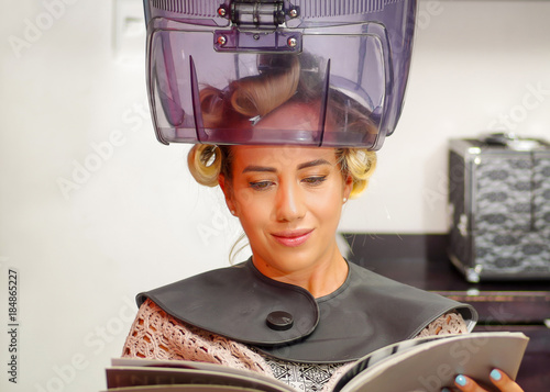 Close up of blonde woman reading a book and waiting under an old hair-dryer chair hood, in a blurred background photo