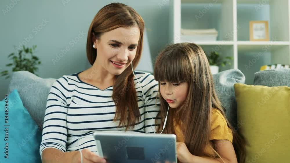Cute little girl with her beautiful mother sitting on the sofa with pillows with headphones, holding tablet computer and laughing. At home. Indoors Cute little girl with her beautiful mother sitting on the sofa with pillows with headphones, holding tablet computer and laughing. At home. Indoors