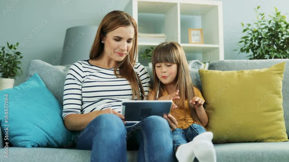 Little girl sitting with her charming mother in headphones listening to the music or watching musical video on the tablet computer at home. Indoor Little girl sitting with her charming mother in headphones listening to the music or watching musical video on the tablet computer at home. Indoor