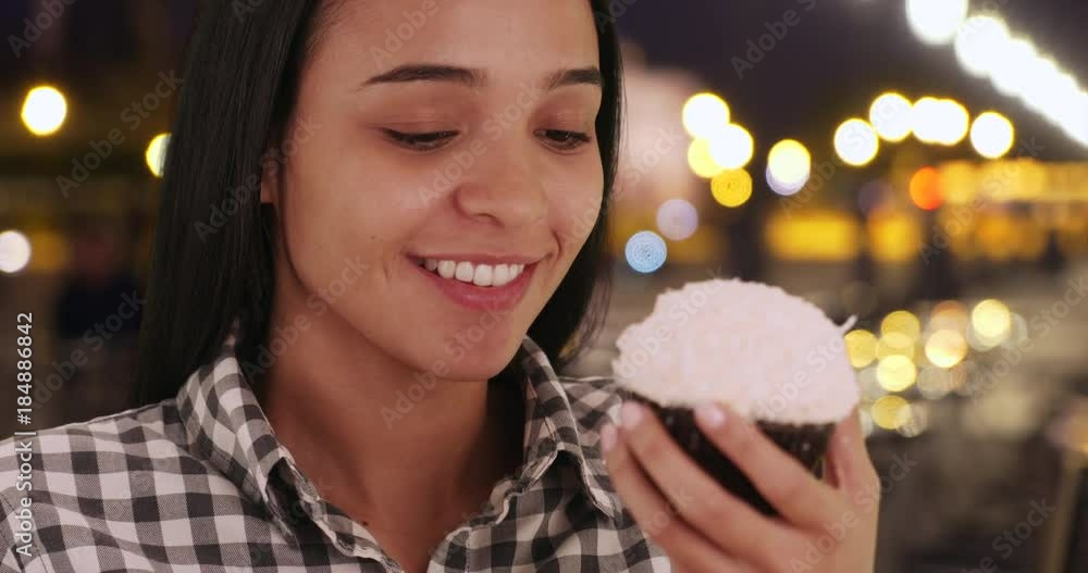 Pretty Hispanic woman savors a freshly baked cupcake in a city street at night. Latina eats a white frosted cupcake on a night out in a brightly lit city. 4k Pretty Hispanic woman savors a freshly baked cupcake in a city street at night. Latina eats a white frosted cupcake on a night out in a brightly lit city. 4k