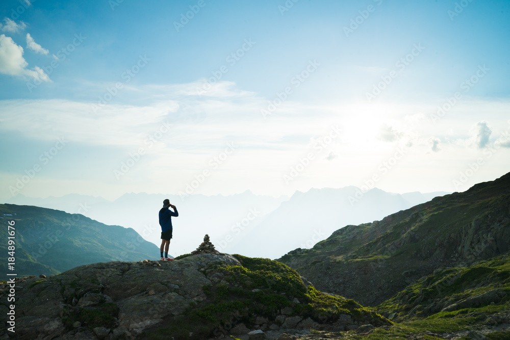 A single hiker silhouetted against a cloudy sky, having a drink at the top of a mountain with mountains and peaks in the distance.