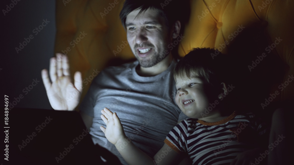 Loving father and his little son having online video chat with grandparents using tablet computer and while lying in bed in evening at home Loving father and his little son having online video chat with grandparents using tablet computer and while lying in bed in evening at home