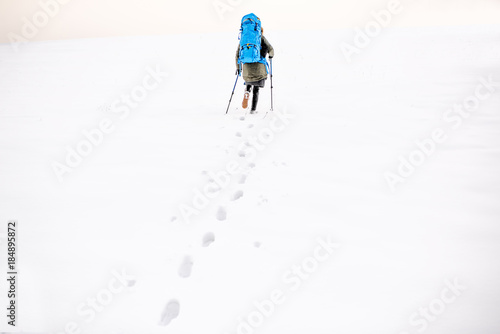 Wide view on the snowy hill with footprints and hiker walking up with backpack in the mountains